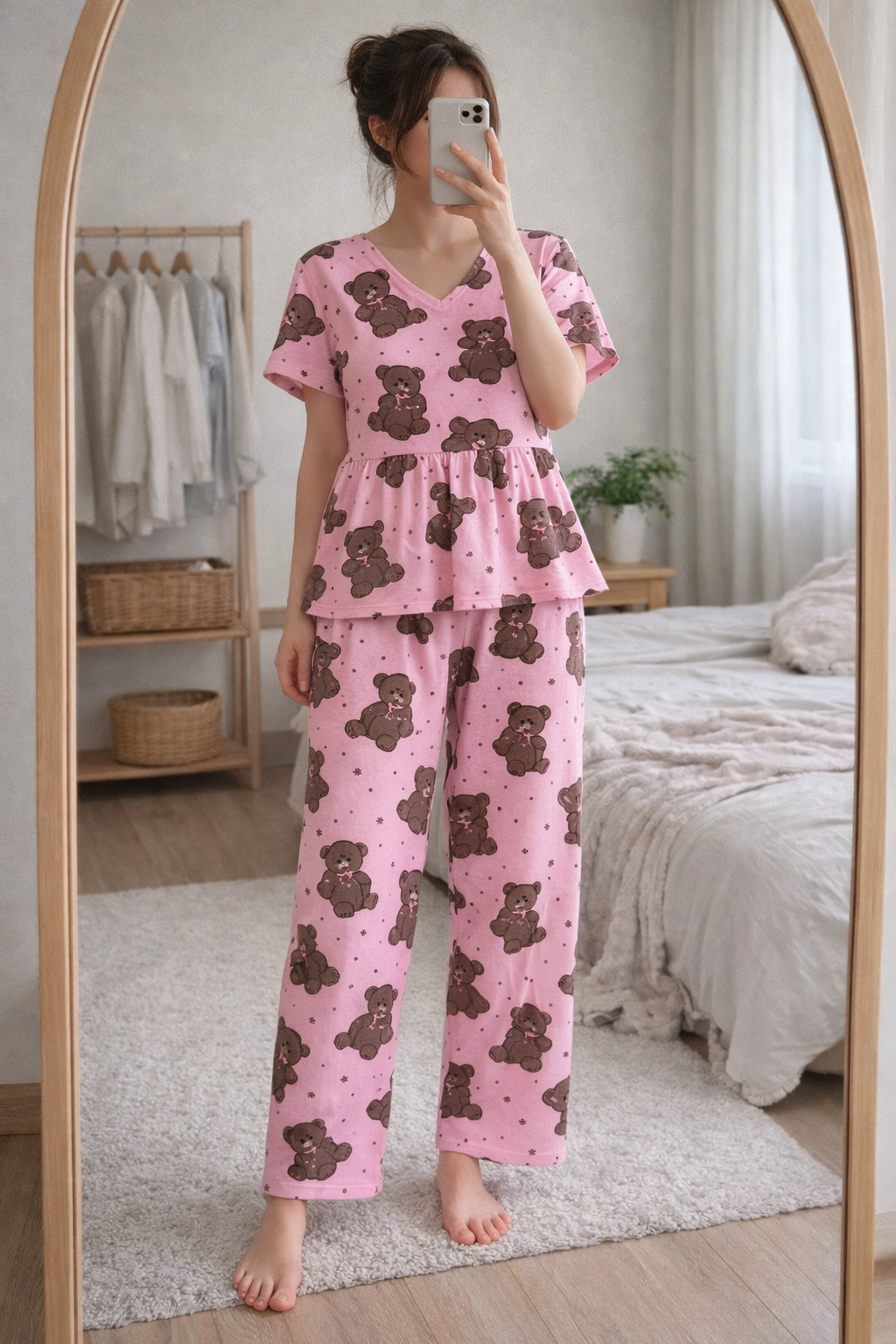 Woman wearing Blush Whispers pink teddy bear print peplum night suit with full-length pants, taking a mirror selfie in a cozy bedroom.