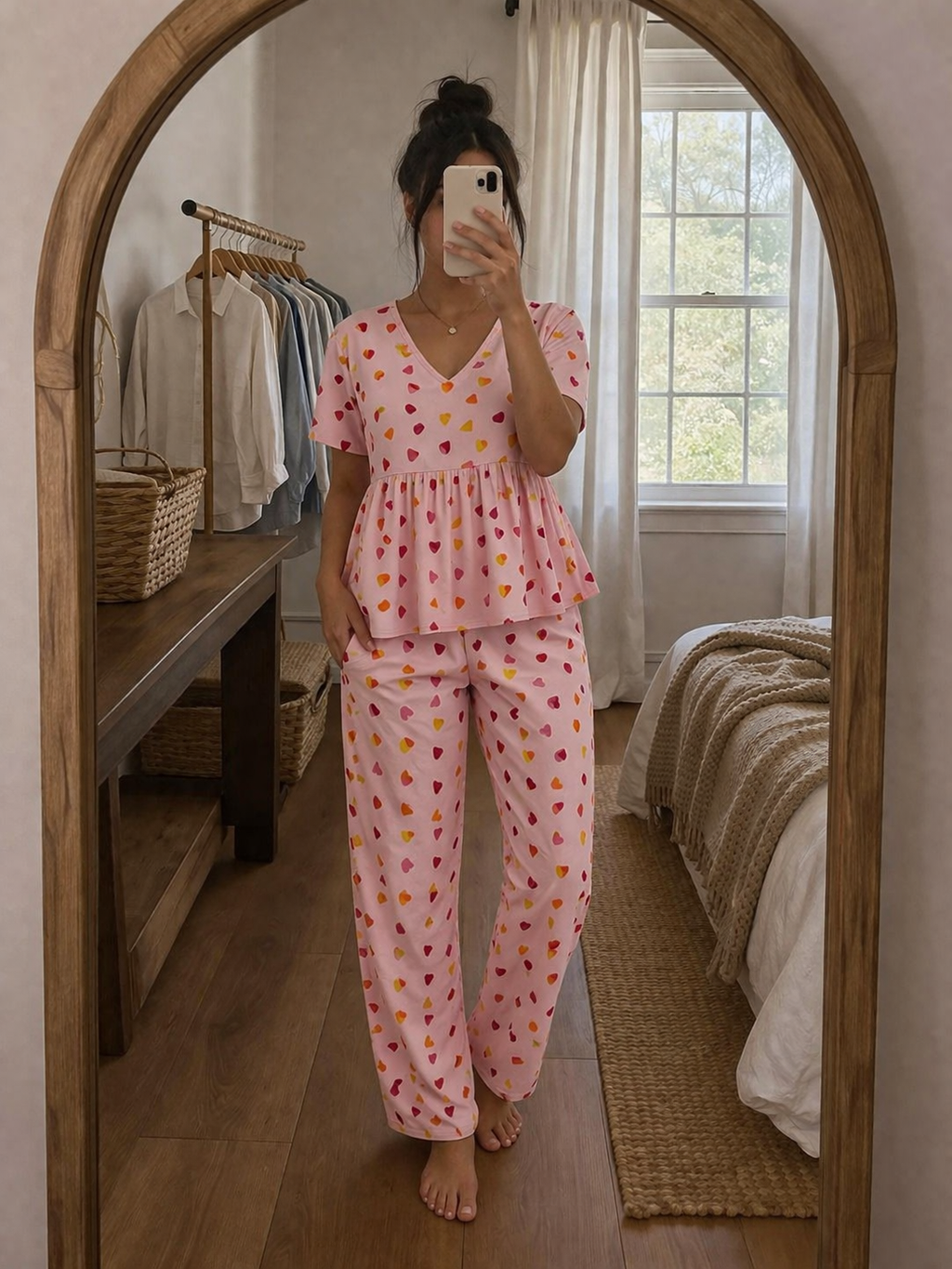 Woman wearing Blush Whispers pink peplum night suit with multicolor heart prints and full-length pants, taking a mirror selfie in a cozy bedroom.