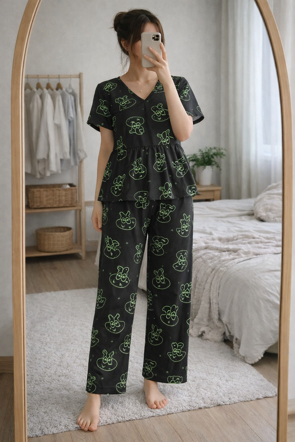 Woman wearing Blush Whispers black peplum night suit with neon green bunny prints and full-length pants, taking a mirror selfie in a cozy bedroom.
