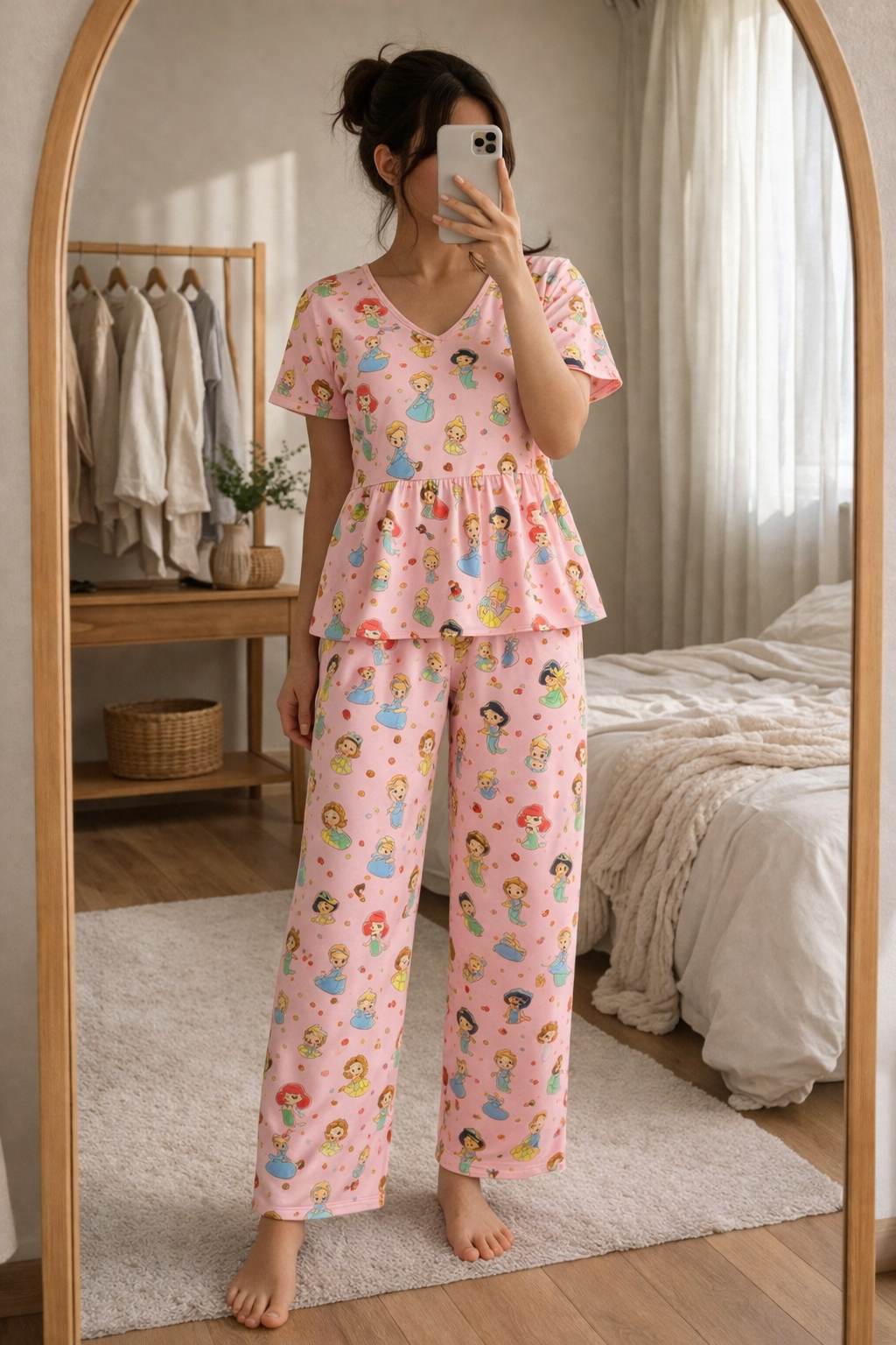 Woman wearing Blush Whispers pink peplum night suit with princess prints and full-length pants, taking a mirror selfie in a cozy bedroom.